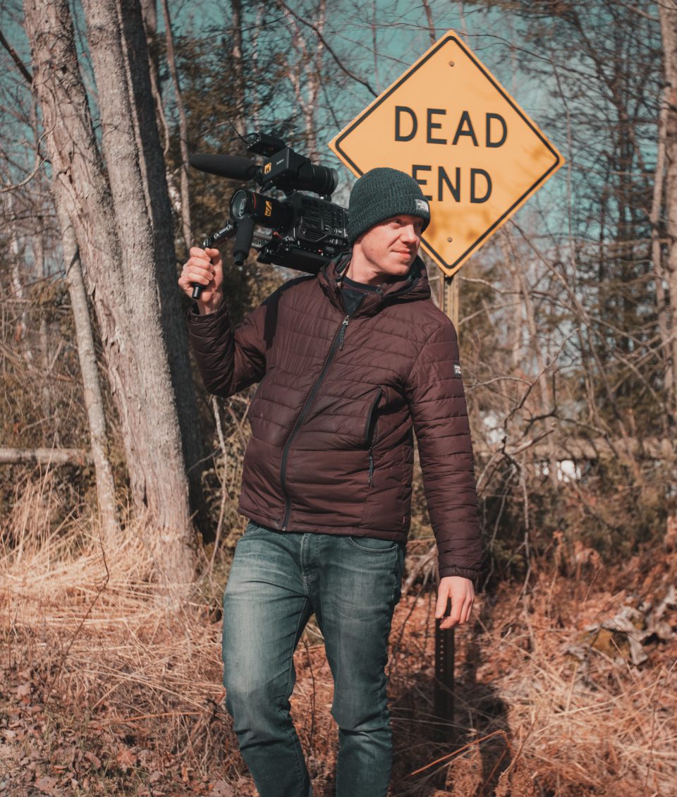 A filmmaker carrying a shoulder-mounted cinema camera on location, beside a yellow Dead End road sign in bare woodland.